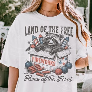 May include: White t-shirt with a graphic featuring a raccoon in a fireworks box, with text reading "LAND OF THE FREE" and "Home of the Feral". The design includes red, white, and blue fireworks and stars.