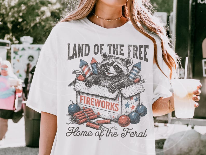 May include: White t-shirt with a graphic featuring a raccoon in a fireworks box, with text reading "LAND OF THE FREE" and "Home of the Feral". The design includes red, white, and blue fireworks and stars.