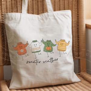 May include: A cream tote bag featuring a playful design of four cartoon sweaters in various colours, holding hands. The words "sweater weather" are written beneath in a handwritten style. The bag has two handles and is made of a light-colored fabric.