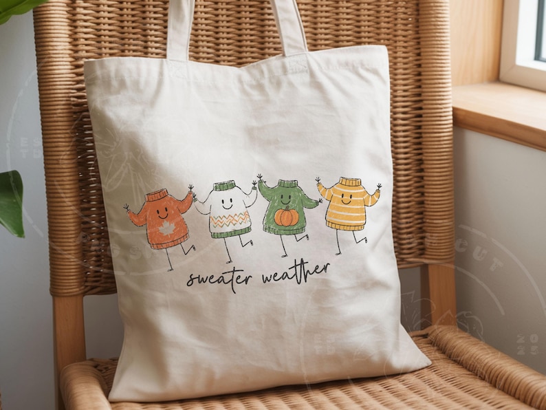 May include: A cream tote bag featuring a playful design of four cartoon sweaters in various colours, holding hands. The words "sweater weather" are written beneath in a handwritten style. The bag has two handles and is made of a light-colored fabric.