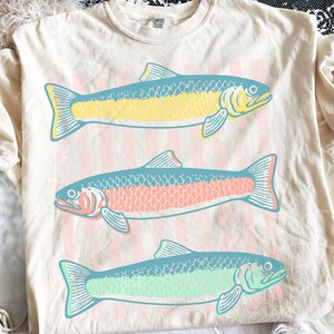 May include: Cream-colored t-shirt featuring three stylized fish in yellow, pink, and green with blue accents. The fish are arranged vertically on a background of pink and white stripes. The shirt has a crew neck and short sleeves.