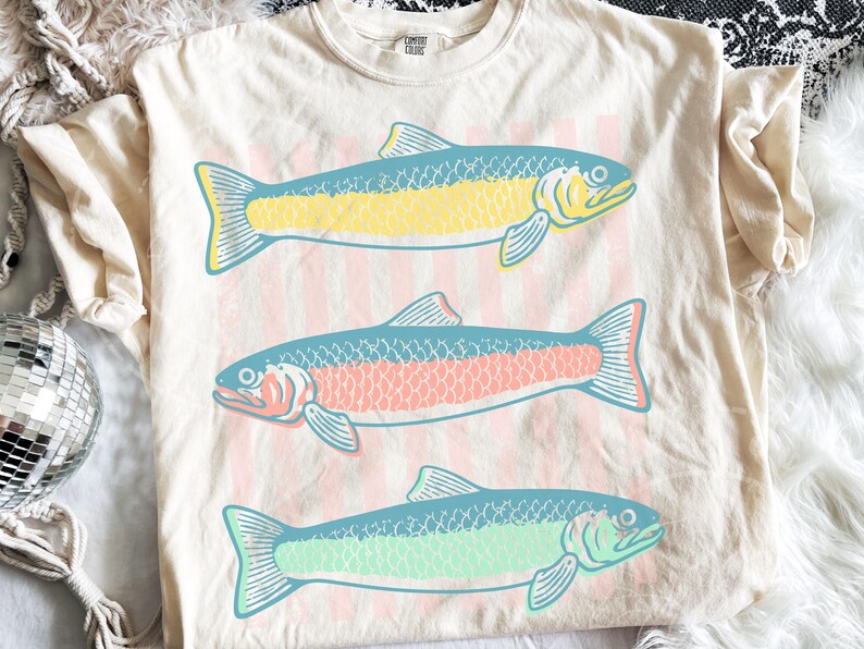May include: Cream-colored t-shirt featuring three stylized fish in yellow, pink, and green with blue accents. The fish are arranged vertically on a background of pink and white stripes. The shirt has a crew neck and short sleeves.
