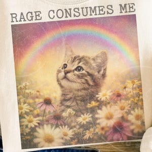 May include: Cream-colored sweatshirt featuring a graphic of a kitten gazing upwards under a rainbow. The kitten is surrounded by flowers, with the text "RAGE CONSUMES ME" above the image. The design has a vintage, distressed look.