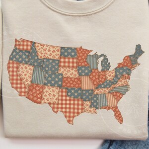 May include: A beige sweatshirt featuring a patchwork design of the United States map. The map is composed of various fabric patterns in red, blue, and white, including stars, stripes, and gingham. The sweatshirt is a casual style.