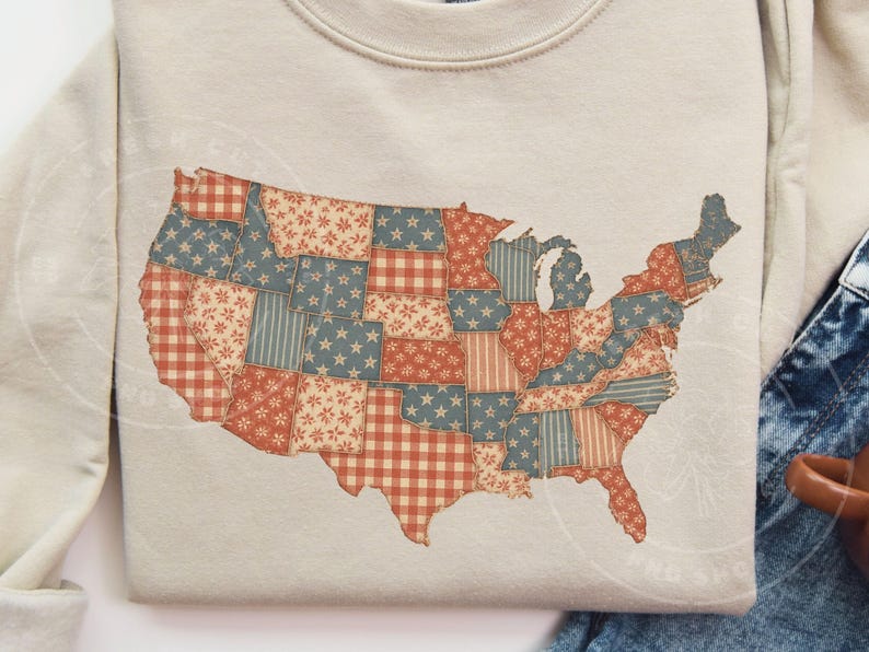 May include: A beige sweatshirt featuring a patchwork design of the United States map. The map is composed of various fabric patterns in red, blue, and white, including stars, stripes, and gingham. The sweatshirt is a casual style.