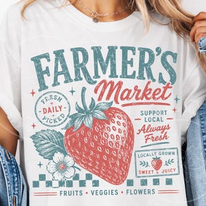 May include: White t-shirt with a vintage-style Farmer's Market graphic. The design features a large red strawberry, text that reads "Farmer's Market," and other supporting text. The shirt promotes fresh, locally grown fruits, veggies, and flowers.