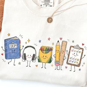 May include: A cream-colored t-shirt featuring a colorful graphic of anthropomorphic school supplies including a blue binder labeled IEP, headphones, a yellow crayon box, a pencil, a ruler, and a clipboard with checkmarks, surrounded by small stars and hearts.