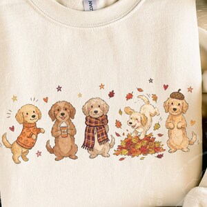 May include: A cream-colored crewneck sweatshirt featuring a graphic of five golden-colored dogs. The dogs are depicted wearing a pumpkin-patterned sweater, holding a coffee cup, wearing a plaid scarf, playing in autumn leaves, and wearing an acorn hat.