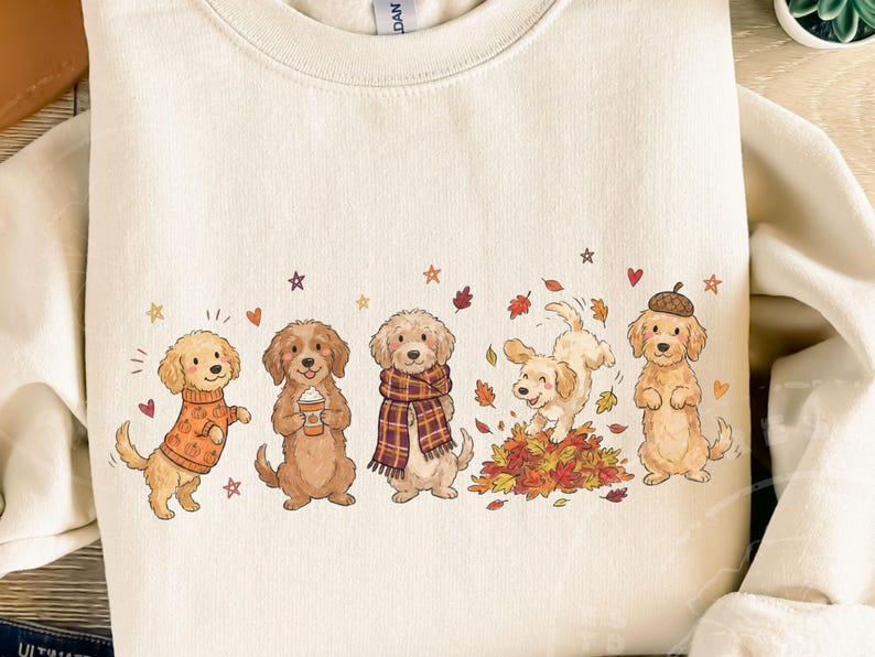 May include: A cream-colored crewneck sweatshirt featuring a graphic of five golden-colored dogs. The dogs are depicted wearing a pumpkin-patterned sweater, holding a coffee cup, wearing a plaid scarf, playing in autumn leaves, and wearing an acorn hat.