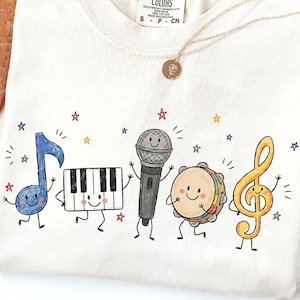 Può includere: Una t-shirt color crema con una stampa grafica di cinque personaggi musicali antropomorfi: una croma blu, una tastiera di pianoforte, un microfono grigio, un tamburello e una chiave di violino gialla, circondati da piccole stelle colorate.