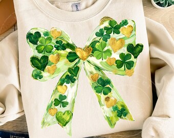 Peut inclure: Sweat-shirt crème avec un motif de nœud peint. Le nœud est orné de trèfles verts, de cœurs verts et d'accents dorés en forme de cœur. Le motif est centré sur le devant du vêtement.