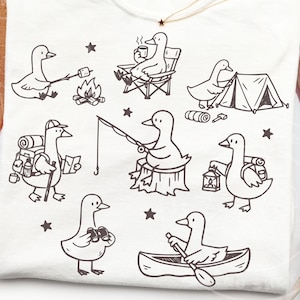 Könnte beinhalten: Weißes T-Shirt mit einer schwarzen Strichzeichnung von Enten beim Camping. Die Enten werden mit Lagerfeuer, Angeln, Zeltaufbau und Kanufahren dargestellt. Das Design umfasst Sterne und Campingausrüstung.