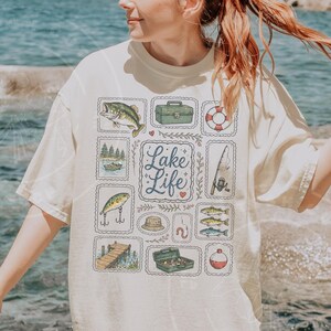 May include: Cream-colored t-shirt featuring a "Lake Life" design. The design includes illustrations of fish, fishing gear, and a lake scene. The shirt has a relaxed fit and short sleeves.