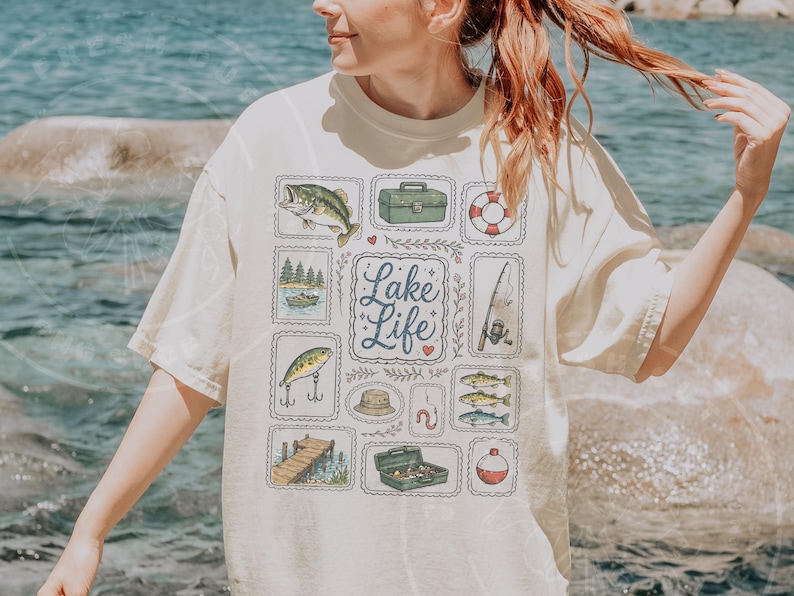 May include: Cream-colored t-shirt featuring a "Lake Life" design. The design includes illustrations of fish, fishing gear, and a lake scene. The shirt has a relaxed fit and short sleeves.