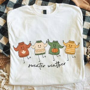 May include: White t-shirt with a playful design of four cartoon sweaters in autumnal hues: rust, cream, green, and yellow. Each sweater has a smiling face and is holding hands. The words "sweater weather" are printed beneath the sweaters.