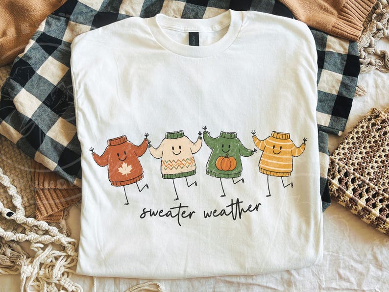 May include: White t-shirt with a playful design of four cartoon sweaters in autumnal hues: rust, cream, green, and yellow. Each sweater has a smiling face and is holding hands. The words "sweater weather" are printed beneath the sweaters.