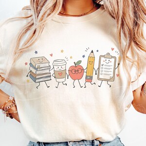 Peut inclure: Un t-shirt couleur cr&egrave;me arborant une impression graphique de cinq fournitures scolaires anthropomorphes se tenant par la main. Les personnages incluent une pile de livres, un gobelet &agrave; caf&eacute;, une pomme &agrave; lunettes, un crayon jaune et un presse-papiers.