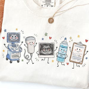 Puede incluir: Una camiseta de color crema con un gráfico colorido de cinco objetos médicos antropomórficos: una máquina de ultrasonido, un transductor, una ecografía, una botella con la etiqueta ULTRASOUND GEL y un portapapeles, todos con caras sonrientes.