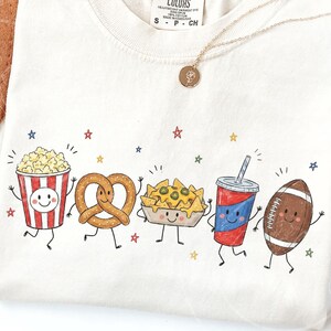 May include: A cream-colored t-shirt featuring a graphic print of five anthropomorphic characters: a popcorn bucket, a pretzel, a tray of nachos, a soda cup, and a football. Each character has a smiling face, arms, and legs, surrounded by small colorful stars.