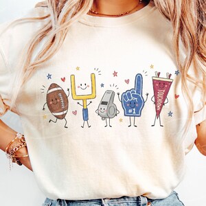 May include: A cream-colored t-shirt featuring a graphic print of five anthropomorphic football-themed characters: a brown football, a yellow goalpost, a grey whistle, a blue foam finger with the number 1, and a maroon pennant labeled TOUCHDOWN.