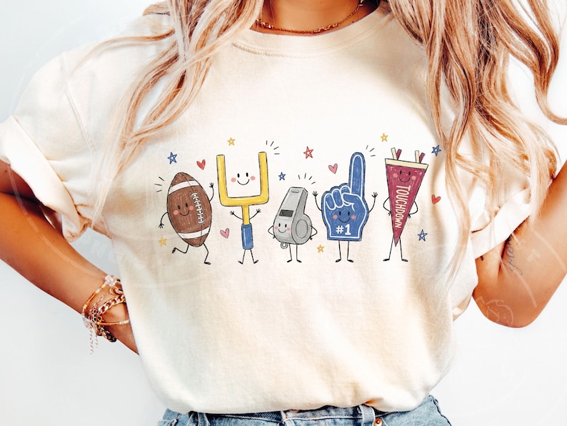 May include: A cream-colored t-shirt featuring a graphic print of five anthropomorphic football-themed characters: a brown football, a yellow goalpost, a grey whistle, a blue foam finger with the number 1, and a maroon pennant labeled TOUCHDOWN.