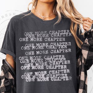 K&ouml;nnte beinhalten: Dunkelgraues T-Shirt mit dem Text "ONE MORE CHAPTER" in verblichener wei&szlig;er Schrift. Der Text wiederholt sich mehrfach auf der Vorderseite. Ein schwarz-wei&szlig; kariertes Hemd wird dar&uuml;ber getragen.