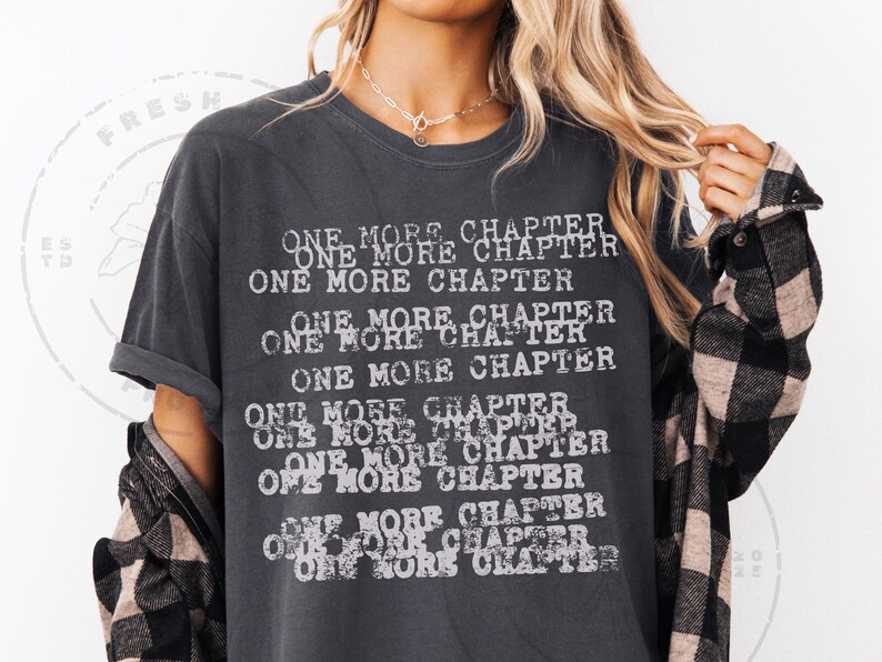 K&ouml;nnte beinhalten: Dunkelgraues T-Shirt mit dem Text "ONE MORE CHAPTER" in verblichener wei&szlig;er Schrift. Der Text wiederholt sich mehrfach auf der Vorderseite. Ein schwarz-wei&szlig; kariertes Hemd wird dar&uuml;ber getragen.