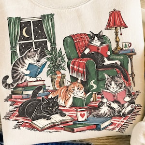 Puede incluir: Una sudadera color crema con una ilustración gráfica de cinco gatos leyendo libros en una habitación acogedora. La escena incluye un sillón verde, una lámpara roja, una ventana con una luna creciente y pilas de libros sobre una alfombra a cuadros.