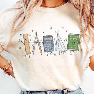 Puede incluir: Una camiseta de color crema con un estampado gr&aacute;fico de cinco &uacute;tiles escolares antropom&oacute;rficos: una regla, un comp&aacute;s, una calculadora, una escuadra y un libro verde con la etiqueta "MATH" con caras sonrientes, rodeados de peque&ntilde;as estrellas coloridas.