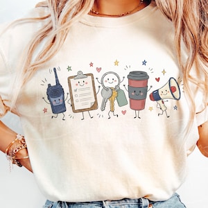 May include: A cream-colored t-shirt featuring a graphic print of five anthropomorphic items: a dark blue walkie-talkie, a tan clipboard, a set of silver and gold keys, a brown coffee cup, and a white megaphone, all accented with small colorful stars and hearts.