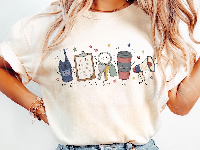 May include: A cream-colored t-shirt featuring a graphic print of five anthropomorphic items: a dark blue walkie-talkie, a tan clipboard, a set of silver and gold keys, a brown coffee cup, and a white megaphone, all accented with small colorful stars and hearts.