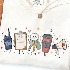 May include: A cream-colored t-shirt featuring a graphic print of five anthropomorphic characters: a dark blue walkie-talkie, a brown clipboard with a checklist, a silver key ring, a red coffee cup, and a white and blue megaphone, all with faces.
