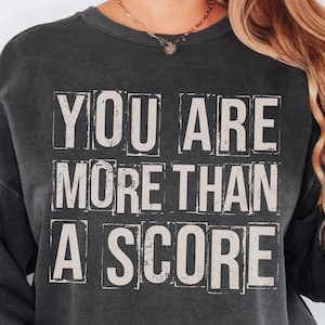 May include: Dark gray sweatshirt with the text "YOU ARE MORE THAN A SCORE" in a distressed, block letter style. The text is in a light beige color, creating a contrast against the dark background. The sweatshirt has a casual, relaxed fit.