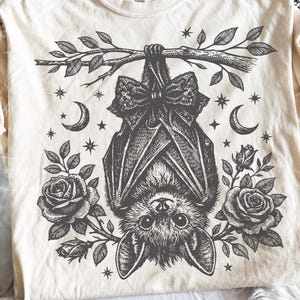 May include: A cream-colored t-shirt featuring a black ink-style graphic of a bat hanging upside down from a leafy branch. The bat wears a large bow, flanked by two blooming roses, crescent moons, and small stars in a detailed, symmetrical design.