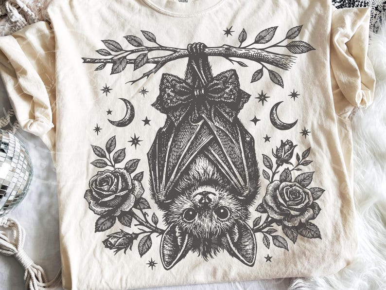 May include: A cream-colored t-shirt featuring a black ink-style graphic of a bat hanging upside down from a leafy branch. The bat wears a large bow, flanked by two blooming roses, crescent moons, and small stars in a detailed, symmetrical design.