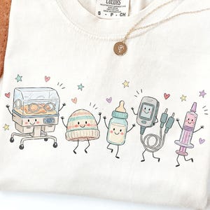 May include: A cream-coloured t-shirt featuring a graphic illustration of five anthropomorphic medical items: a neonatal incubator, a striped baby hat, a baby bottle, a pulse oximeter, and a syringe, all surrounded by small colourful hearts and stars.