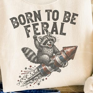 May include: Cream-colored sweatshirt with the text "BORN TO BE FERAL" above a raccoon riding a rocket. The raccoon is gray and white, and the rocket is red, white, and blue. Stars and streaks of smoke emanate from the rocket.