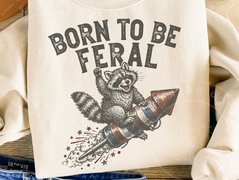 May include: Cream-colored sweatshirt with the text "BORN TO BE FERAL" above a raccoon riding a rocket. The raccoon is gray and white, and the rocket is red, white, and blue. Stars and streaks of smoke emanate from the rocket.
