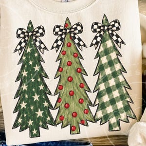 Könnte beinhalten: Ein cremefarbenes Sweatshirt mit Rundhalsausschnitt und einem Grafikdruck von drei stilisierten Weihnachtsbäumen. Jeder Baum ist mit einer schwarz-weiß karierten Schleife verziert. Der erste Baum hat ein Sternenmuster, der zweite rote Kugeln und der dritte ein grün-weißes Karomuster.