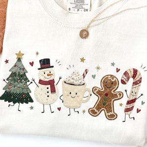 May include: A cream-colored t-shirt featuring a festive graphic print of a decorated Christmas tree, a snowman with a red scarf, a mug of hot cocoa with whipped cream, a gingerbread person, and a candy cane, all with small arms and legs.