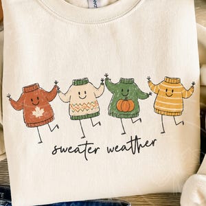 May include: A cream-coloured sweatshirt with a playful design of four animated sweaters in autumnal hues: rust, cream, green, and yellow. Each sweater has a smiling face and is dancing. The words "sweater weather" are written in a script font below.