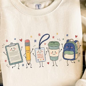 May include: A cream-colored crewneck sweatshirt featuring a graphic print of five anthropomorphic school supplies: a clipboard, a yellow pencil, a hall pass badge, a green coffee cup, and a blue backpack, all accented with small colorful stars.