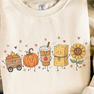 Könnte beinhalten: Ein cremefarbenes Sweatshirt mit Rundhalsausschnitt und einem Grafikdruck von fünf lächelnden herbstlichen Charakteren: ein mit Kürbissen gefüllter Holzwagen, ein großer orangefarbener Kürbis, ein Kaffeebecher, ein Heuballen und eine gelbe Sonnenblume.