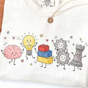 May include: A cream-colored t-shirt featuring a graphic print of five anthropomorphic characters: a pink brain, a yellow lightbulb, a stack of red, blue, and yellow building blocks, two grey gears, and a grey chess rook, all with smiling faces.