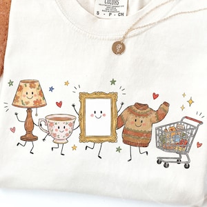 Könnte beinhalten: Ein cremefarbenes T-Shirt mit einem Grafikdruck von fünf anthropomorphen Objekten: einer Blumenlampe, einer Teetasse, einem goldgerahmten Spiegel, einem gemusterten Pullover und einem Einkaufswagen, alle mit lächelnden Gesichtern und kleinen Sternen und Herzen.