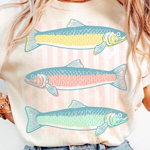 May include: Cream-colored t-shirt featuring three stylized fish in shades of yellow, pink, and green. The fish are set against a backdrop of vertical pink and white stripes. The design is simple and eye-catching, perfect for casual wear.