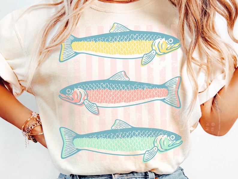 May include: Cream-colored t-shirt featuring three stylized fish in shades of yellow, pink, and green. The fish are set against a backdrop of vertical pink and white stripes. The design is simple and eye-catching, perfect for casual wear.