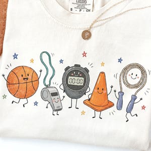 May include: A cream-colored t-shirt featuring a graphic print of five anthropomorphic sports items: an orange basketball, a grey whistle, a black digital stopwatch displaying 00:00, an orange traffic cone, and a jump rope with blue handles.
