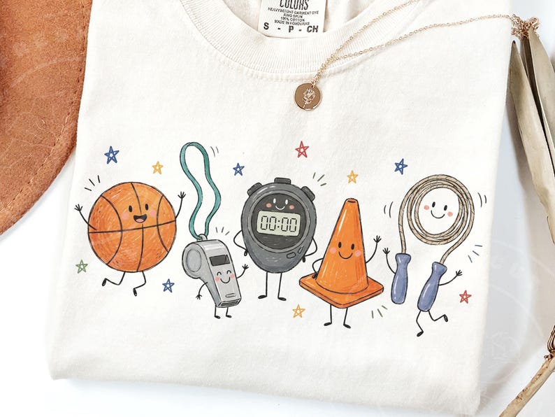 May include: A cream-colored t-shirt featuring a graphic print of five anthropomorphic sports items: an orange basketball, a grey whistle, a black digital stopwatch displaying 00:00, an orange traffic cone, and a jump rope with blue handles.