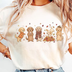 May include: A cream-colored t-shirt featuring a graphic print of five golden-colored dogs in various autumn-themed poses. The dogs are depicted wearing sweaters, scarves, and an acorn hat, with some playing in a pile of colorful fall leaves.